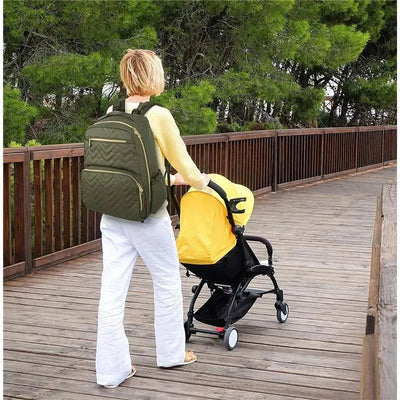 Fisher Price Morgan Diaper Bag Backpack in olive carried by a person on a wooden path beside a stroller with a yellow hood.