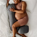 Pregnant person resting on a white bed, embracing a gray Frida Mom adjustable pregnancy body pillow.