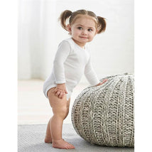 White long-sleeve baby bodysuit worn by a toddler with pigtails, standing near a knitted ottoman in a bright setting.