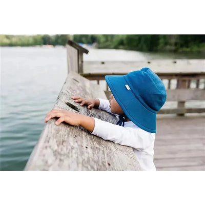 Baby wearing a navy Green Sprouts breathable swim and sun bucket hat, leaning over a wooden railing by a lake.