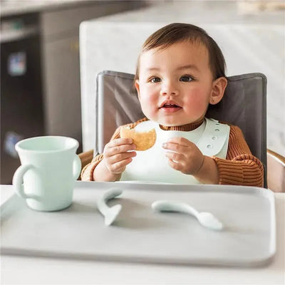 Light blueberry silicone learning cup next to a smiling toddler in a high chair, holding a snack.
