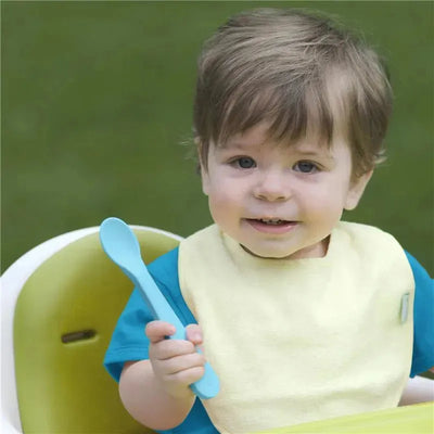 Infant boy wearing a yellow bib, holding a blue spoon, seated in a high chair, smiling outdoors.