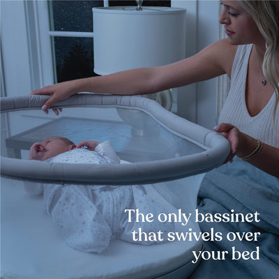 HALO BassiNest Soothing Swivel Sleeper in grey, featuring a baby resting inside and a caregiver adjusting it by the mesh side.