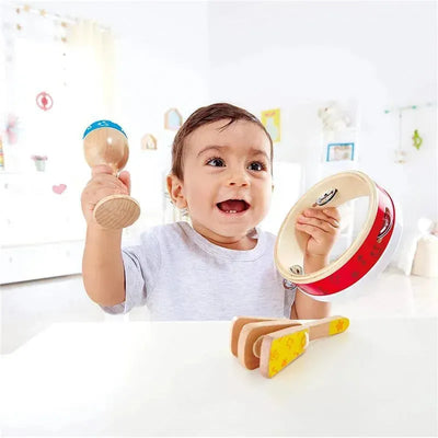A cheerful toddler holding a maraca and a tambourine, with wooden rhythm instruments on a table.