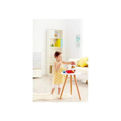 Child in a yellow dress using a toy smoothie blender at a small table with colorful play food in a bright room.