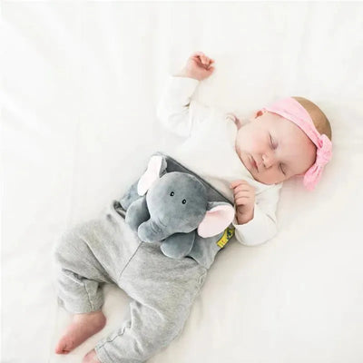 Baby sleeping peacefully with a gray elephant plush toy inserted in a waistband, wearing a pink headband.