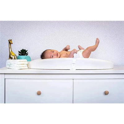 A baby lying on a white Hatch Baby changing pad and scale, with a gold giraffe statue and a potted plant nearby.