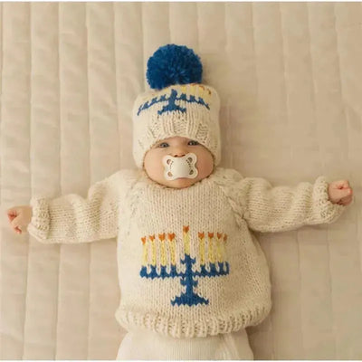 Baby wearing a cream-colored Menorah-themed sweater with a matching hat, laying on a beige surface.