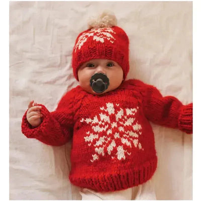 Baby wearing a red snowflake sweater and matching hat, lying on a soft surface with a pacifier.