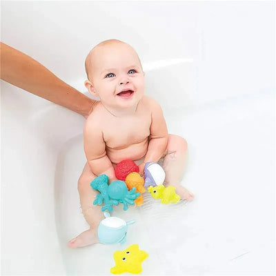Baby sitting in a bathtub surrounded by colorful Infantino aquarium bath squirters, smiling happily while playing.