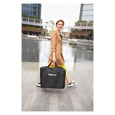 Black Inglesina Quid³ stroller carry bag held by a woman in a tan coat, walking by a reflective fountain.