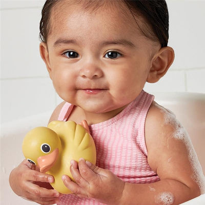 A baby in a pink striped swimsuit smiles while holding a yellow rubber duck in a bubbly bath setting.