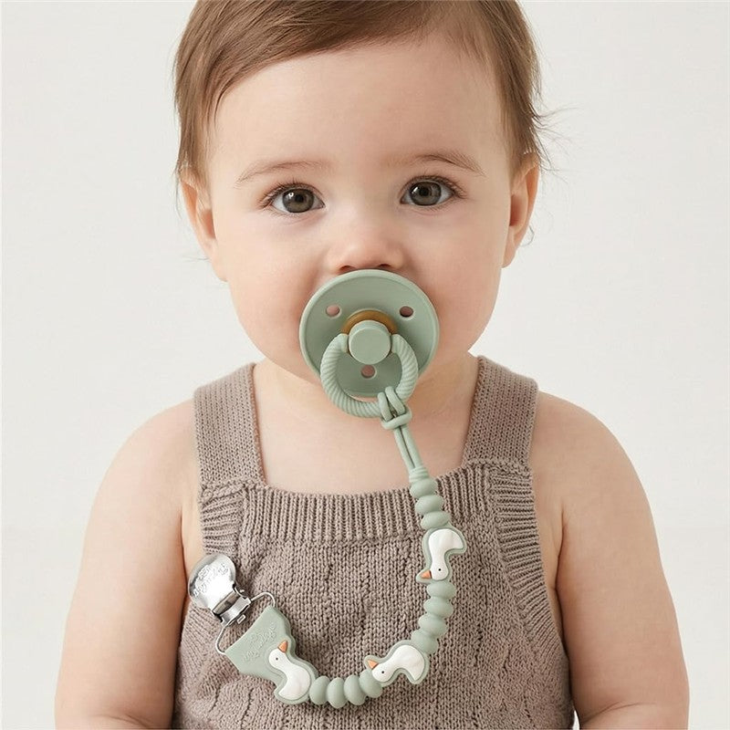 Baby wearing a sage green pacifier attached to a strap featuring goose designs, dressed in a knitted tank top.