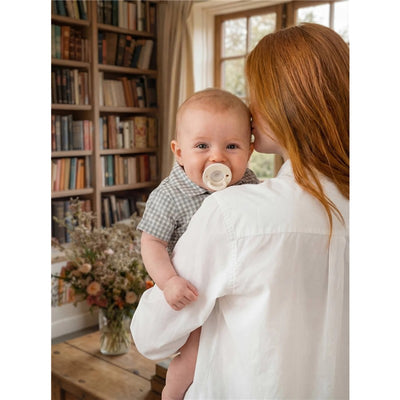 Baby holding an Itzy Ritzy Latch & Soothe pacifier in taupe, smiling at the camera while being cradled by a woman.