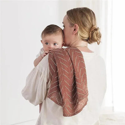 A woman holds a baby while using the Itzy Ritzy Breastfeeding Boss Multi-Use Cover, featuring a brown design with white accents.