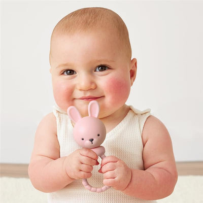 Baby smiles while holding a pink bunny rattle with long ears and a round head, wearing a light-colored outfit.