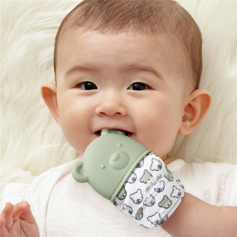 Baby smiling while holding an Itzy Ritzy Sage Bear Teething Mitt, featuring a green bear design with a fabric sleeve.