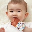 Baby smiling while holding a cow-themed silicone teething mitten, featuring colorful patterns on the fabric.