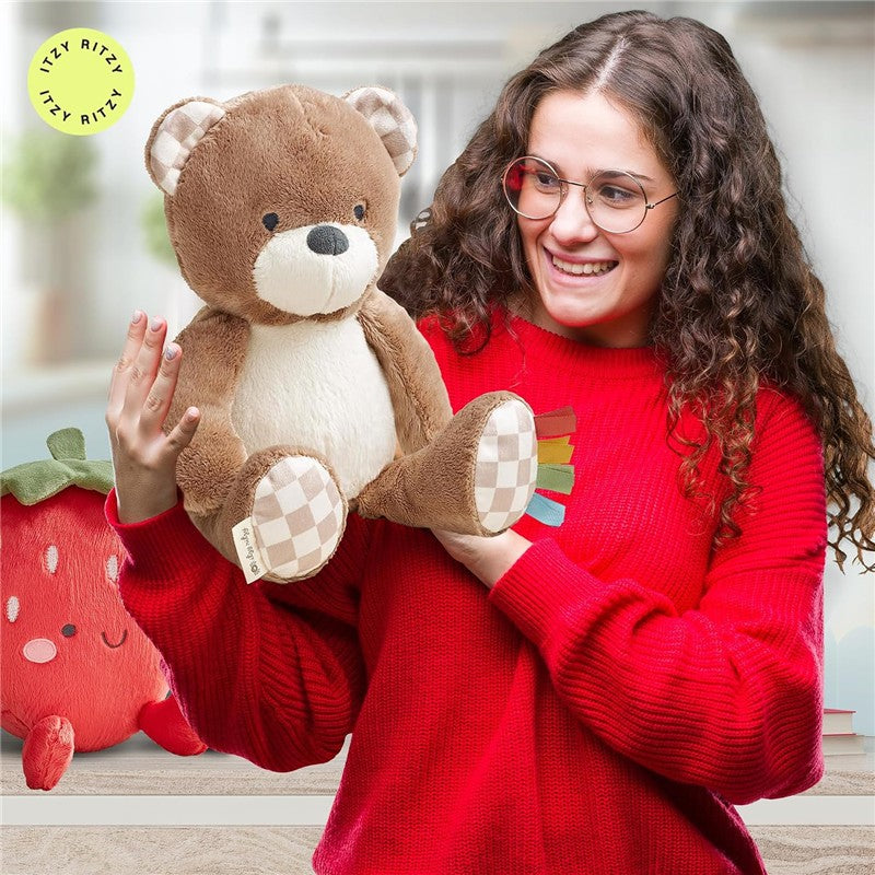 Soft brown bear plush toy with a cream belly, held by a smiling woman in a red sweater; a strawberry toy is visible behind.