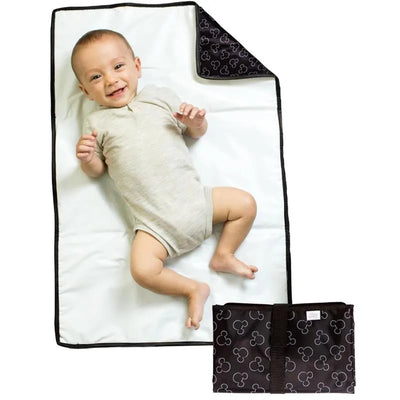 Portable changing pad featuring a white surface and Mickey Mouse patterned black fabric, with a baby laying on it, smiling.