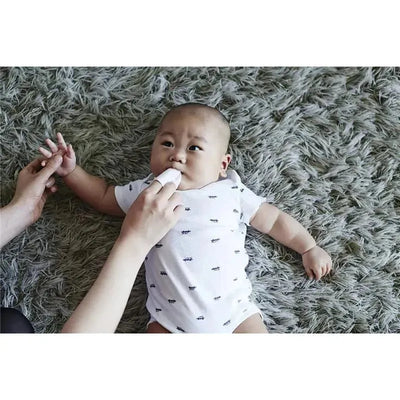 Baby lying on a soft rug, receiving dental care with a silicone finger brush held by an adult.