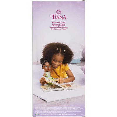 A young girl with curly hair smiles while reading a book, holding a Tiana doll in a green dress.