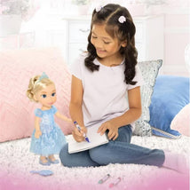 A young girl with curly hair plays with a 14-inch Cinderella doll, drawing in a notebook on a fluffy carpet.