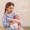 Child with braided hair and a gray dress, surprised while holding a 15.5” reborn baby doll in a pink outfit with a heart design.