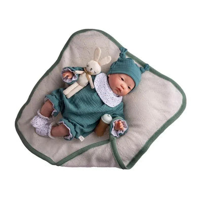 Vinyl boy doll dressed in a blue outfit with a bunny toy, lying on a soft blanket with a bottle nearby.
