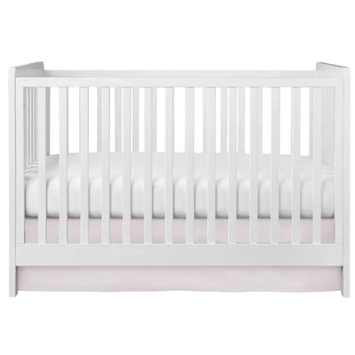 Pink crib skirt underneath a white slatted crib with a white mattress visible inside.