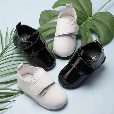 White and black baby boy Oxford shoes with Velcro straps, placed on a light blue background with tropical leaves.