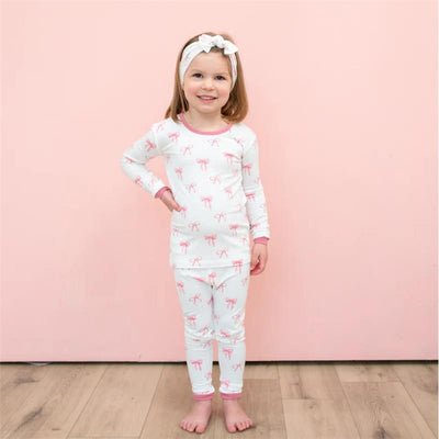 Baby girl wears long sleeve pajamas featuring pink bows, paired with a matching headband, smiling against a pink background.