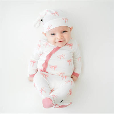 Baby girl wearing a white take-me-home set with pink bow patterns and a matching cap, smiling while seated.