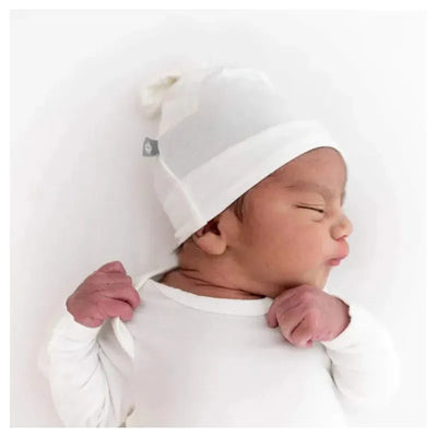Bamboo baby knotted cap in cloud worn by a newborn, lying down and smiling slightly in a white onesie.