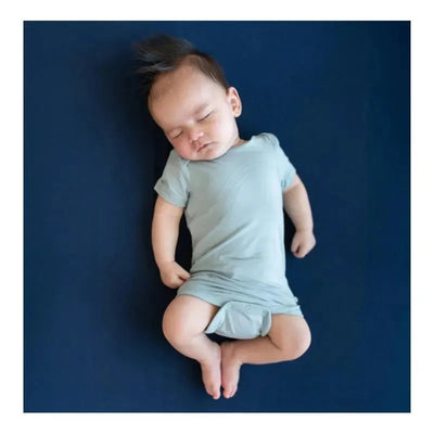 A sleeping baby in a light fog-colored bodysuit rests on a dark blue surface, showcasing its soft fabric and comfort.