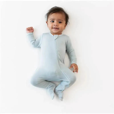 Baby wearing a light gray zippered footie, lying on a white background, smiling and raising one hand.