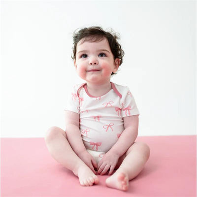 Baby sitting on a pink surface, wearing a white bodysuit with pink bow designs, smiling softly.