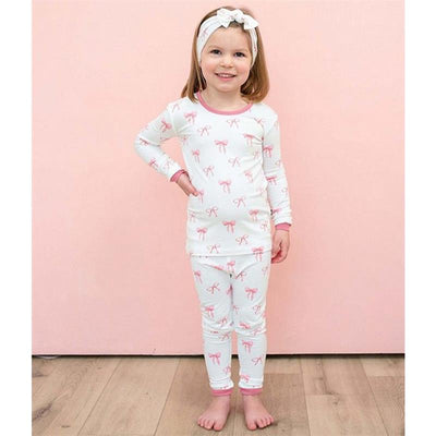 Kyte Baby Long Sleeve Pajamas in Bow for 5T, featuring a young girl wearing white pajamas with pink bow prints and a matching headband.