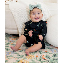 Baby wearing a black floral Kyte Baby long sleeve twirl bodysuit dress, smiling while sitting on a colorful rug, with a green headband.