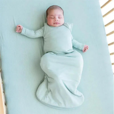 Bamboo sleep bag in sage color, worn by a peacefully sleeping baby, lying on a light blue crib mattress.
