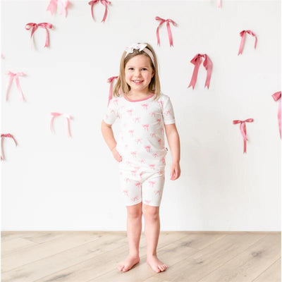 Short sleeve pajamas for kids featuring a bow print, worn by a smiling girl against a backdrop of pink ribbons.