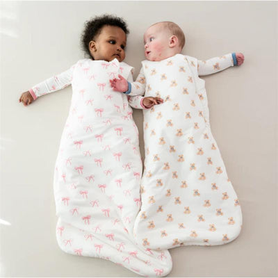 Two babies resting on a light surface, each in a Kyte Baby Sleep Bag; one features pink bows, the other has teddy bear prints.