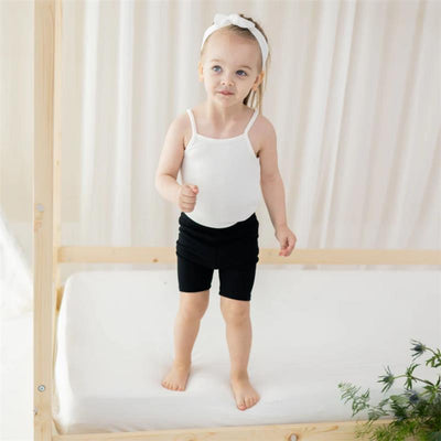 Toddler wearing a white spaghetti strap leotard and black shorts, standing on a bed with wooden frame and soft backdrop.