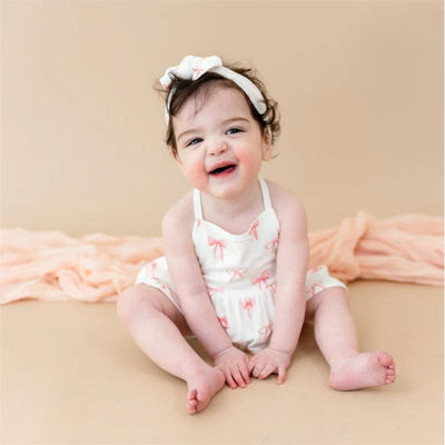 Baby wearing a white spaghetti strap romper with pink bows, sitting and smiling, with a matching headband.
