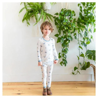 Toddler wearing a Kyte Baby pajama set featuring a light blue dinosaur print, standing in a bright room with plants.
