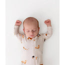 Zippered footie in beige with dachshund prints, worn by a sleeping baby with arms raised, laying on a white surface.