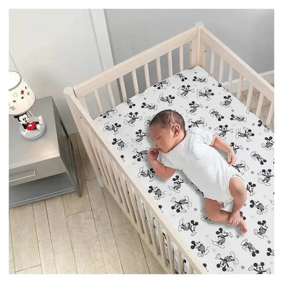 Crib fitted sheet featuring playful Mickey Mouse designs, with a sleeping baby in a white onesie in a wooden crib.