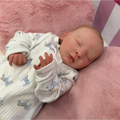Reborn Baby Doll named Joseph, made of white vinyl, wearing a striped outfit, resting on a pink surface with eyes closed.
