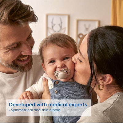 Two smiling parents hold a baby with a beige orthodontic pacifier, showcasing its symmetrical and thin nipple design.