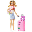 Barbie doll in a casual outfit holds a phone, with a pink suitcase featuring stickers and a purple neck pillow beside her.